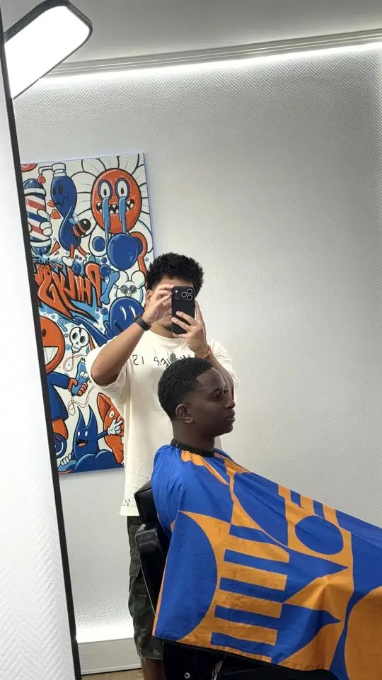 Dans le grand miroir du studio : un client sous la cape, et la fresque bleue et orange au mur.